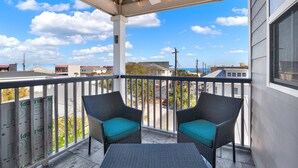 A balcony with two wicker chairs and a table, overlooking a street with buildings and a clear sky. - Beach Baby! Steps to South beach, Pool Access (Tybee Island)
