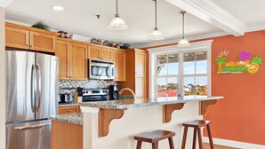 A modern kitchen with wooden cabinets, stainless steel appliances, a granite countertop, and a bar stool. - Beach Baby! Steps to South beach, Pool Access (Tybee Island)