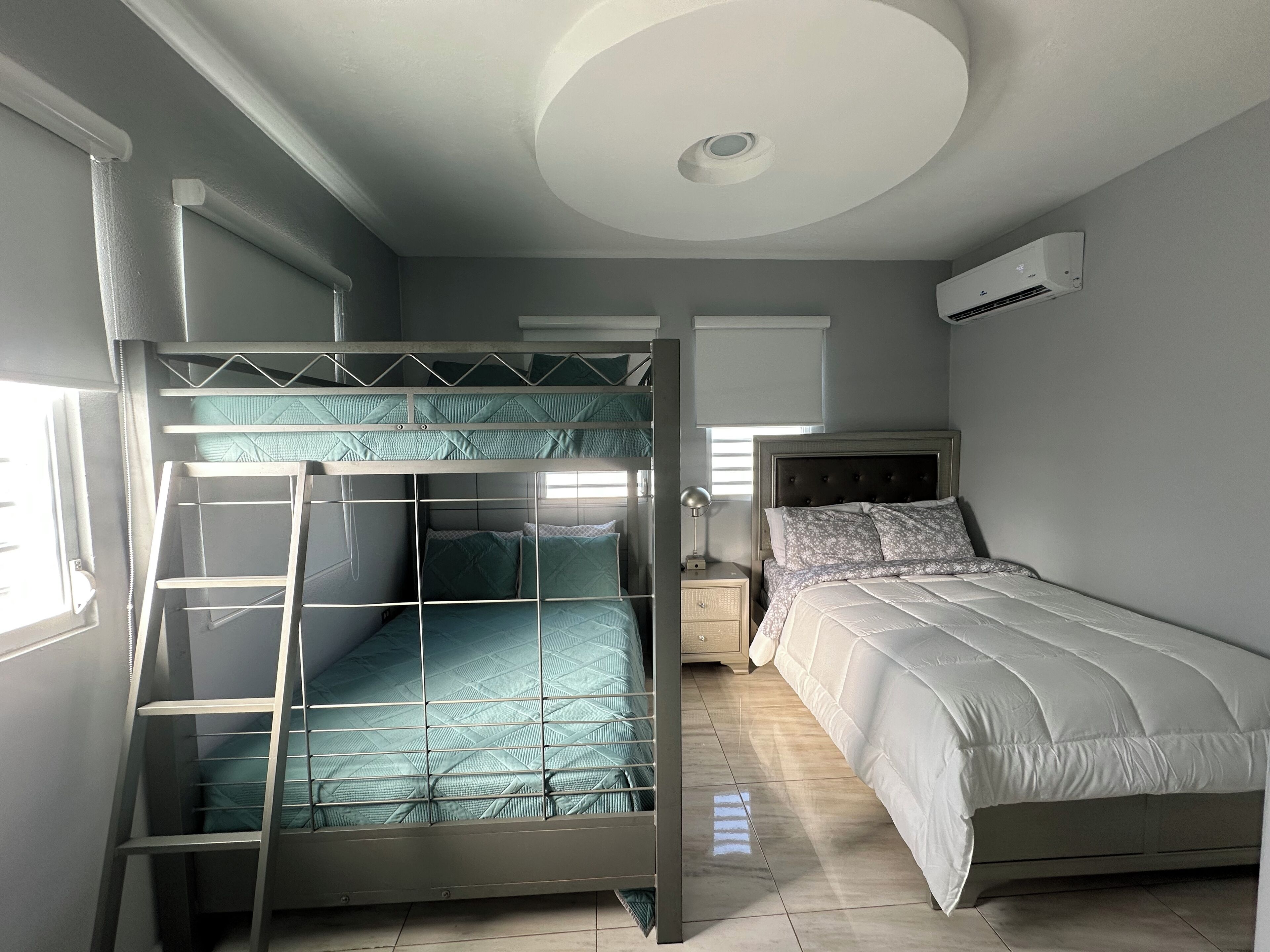 Une chambre avec un lit superposé, un lit simple, une table de chevet, une lampe et un appareil de climatisation.
