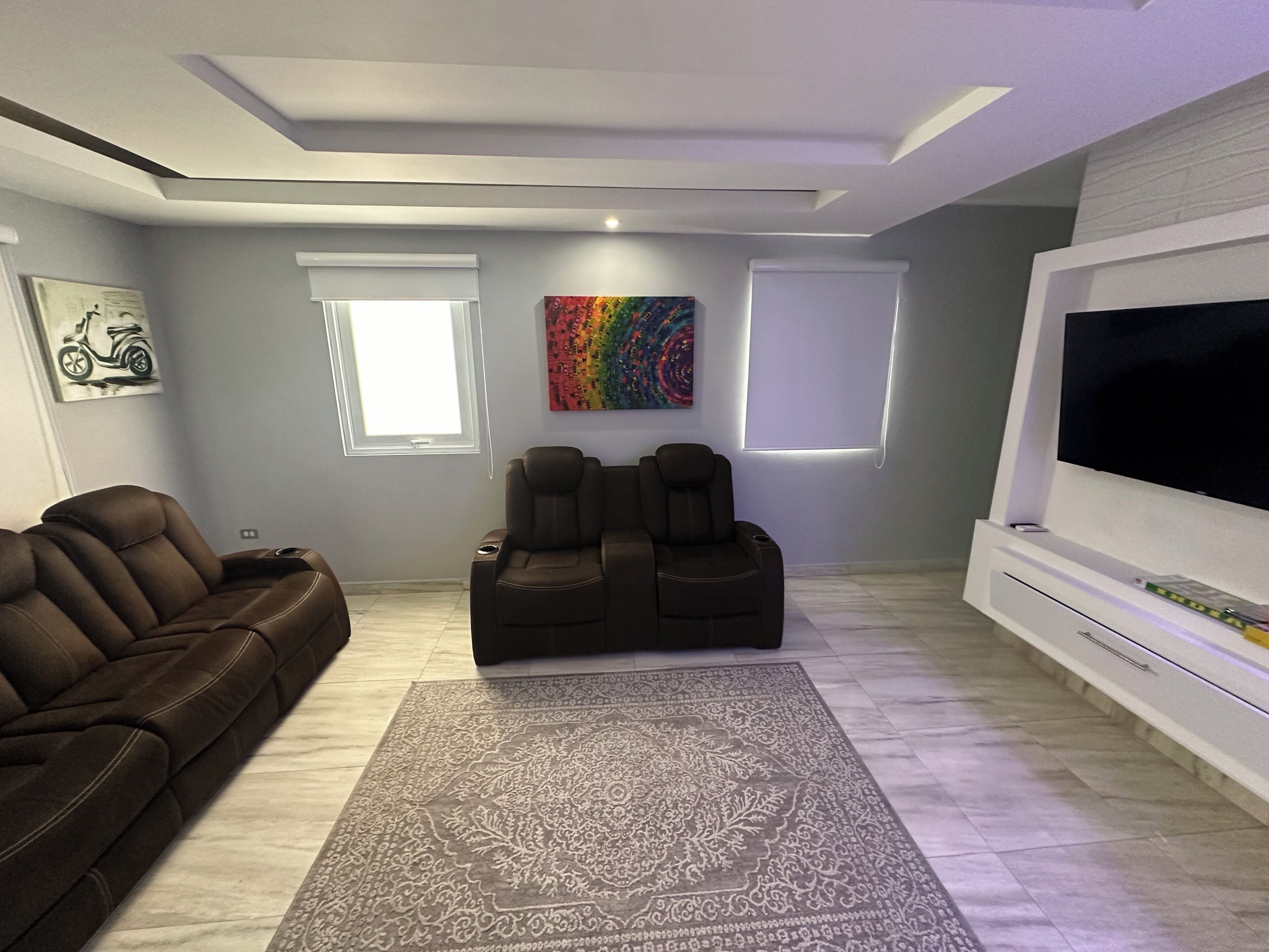 En moderne stue med en brun lædersofa, to lænestole, et mønstret tæppe, et stort fladskærms-TV og kunst på væggene.