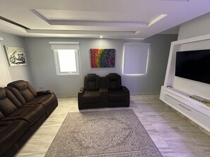 Una sala de estar moderna con un sofá de cuero marrón, dos sillones reclinables, un tapete estampado, un televisor de pantalla plana grande y obras de arte en las paredes.
