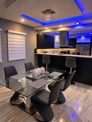A modern dining area with a glass table, black chairs, and a kitchen in the background.