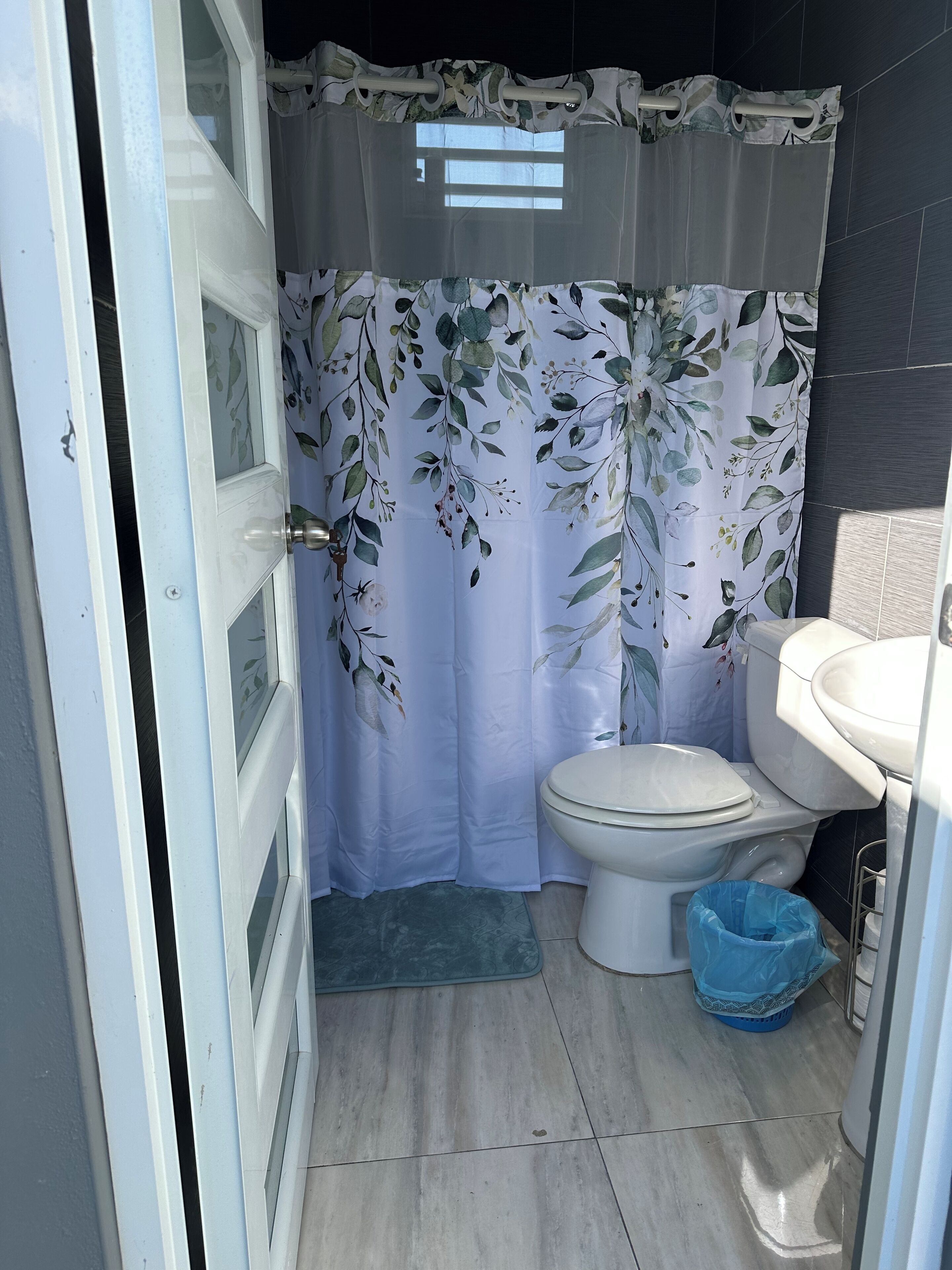 Een badkamer met toilet, wastafel, blauwe prullenbak en een douchegordijn met bloemenmotief.