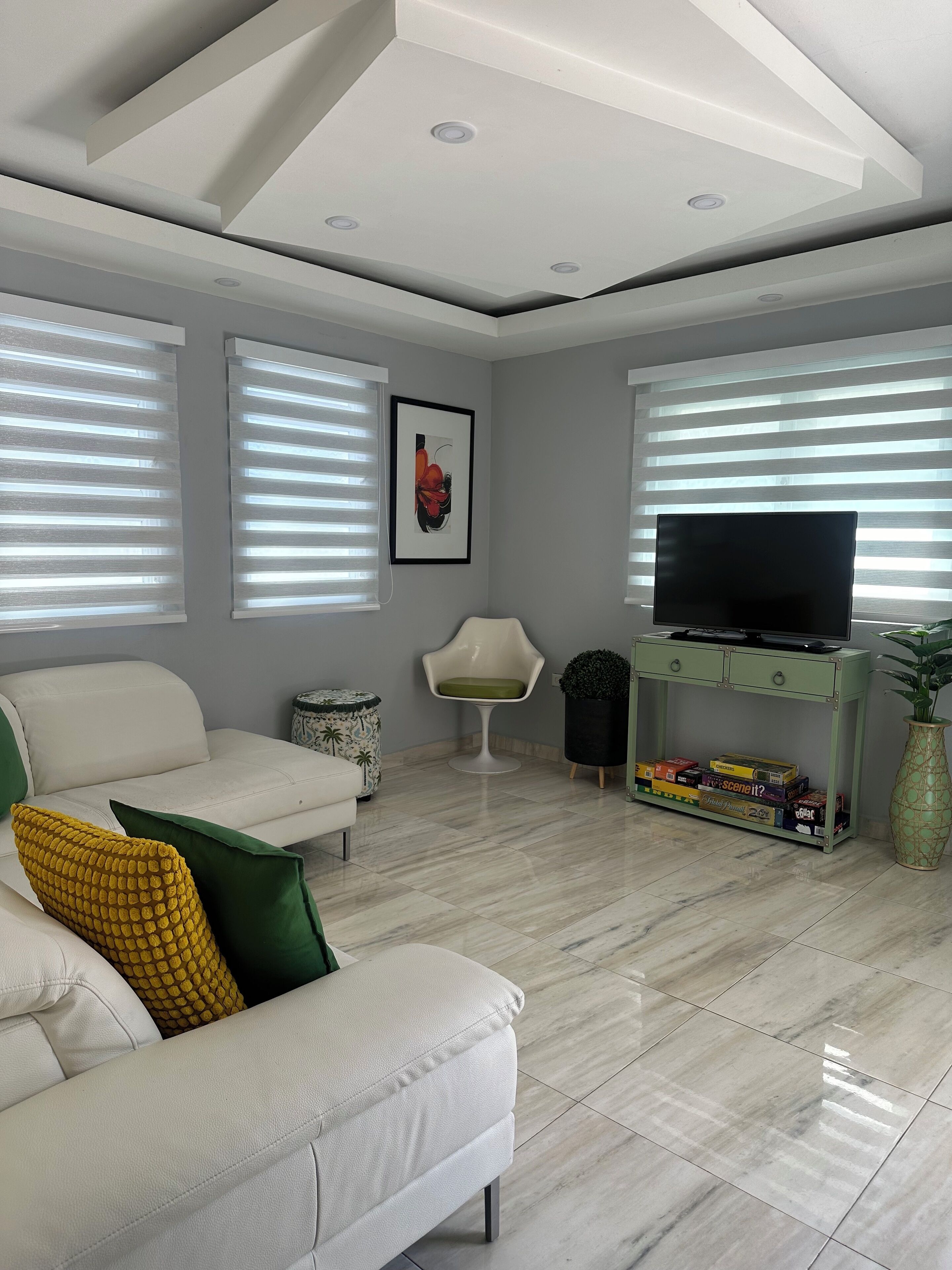 A modern living room with a white sofa, green and yellow cushions, a flat-screen TV, and a green entertainment unit with board games.