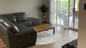 A modern living room with a leather sofa, a wooden coffee table, and a large sliding glass door leading to an outdoor area. - Modern Four Bedroom Holiday Home Available in Deebing Heights (Deebing Heights)