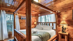 Een slaapkamer in een houten chalet, met een hemelbed, een nachtkastje met lamp en uitzicht op bomen.