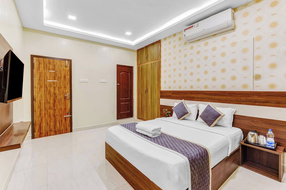 A hotel room with a large bed, a wooden headboard, a bedside table with a kettle and a bottle, and an air conditioning unit on the wall.