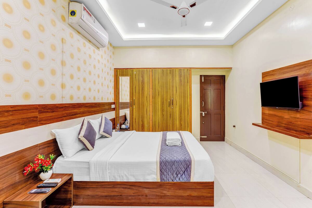 A modern hotel room with a large bed, wooden headboard, a flat-screen TV mounted on the wall, and a ceiling fan.