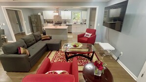 Un salón moderno con un sofá modular, un sillón rojo y una mesa de centro de vidrio. Se ve una cocina al fondo, con armarios blancos y un horno empotrado.