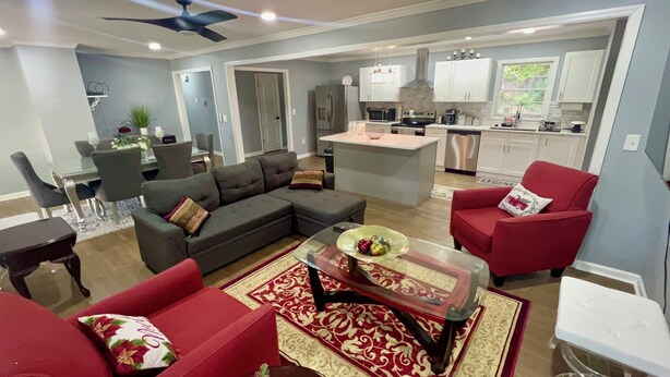 Una sala de estar moderna con un sofá gris, sillones rojos y una mesa de centro de vidrio. Se ve una cocina con armarios blancos y electrodomésticos de acero inoxidable al fondo.
