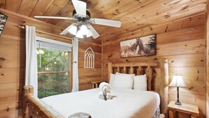 A wooden cabin bedroom with a bed, a ceiling fan, a window with a view of trees, and a wooden nightstand with a lamp. - Seclusion! I Privacy I Fire Pit I Game Room I Arcades I Pet Friendly I Hot Tub (Sevierville)