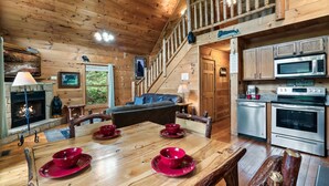 A wooden cabin interior with a dining table set for four, a kitchen with modern appliances, a fireplace, and a staircase leading to a loft area. - Seclusion! I Privacy I Fire Pit I Game Room I Arcades I Pet Friendly I Hot Tub (Sevierville)