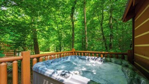 A hot tub surrounded by a wooden deck and dense green trees. - Newly Built-Sleeps 8 w/Fire Pit, Arcade lineup, Hot Tub, Horseshoe Pit & BBall (Sevierville)