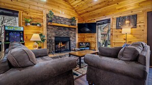 A log cabin interior with a stone fireplace, a television, and a Pac-Man arcade machine. - Newly Built-Sleeps 8 w/Fire Pit, Arcade lineup, Hot Tub, Horseshoe Pit & BBall (Sevierville)