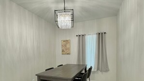 Un comedor con una mesa rectangular, cuatro sillas, una araña, una ventana con cortinas y un letrero en la pared.