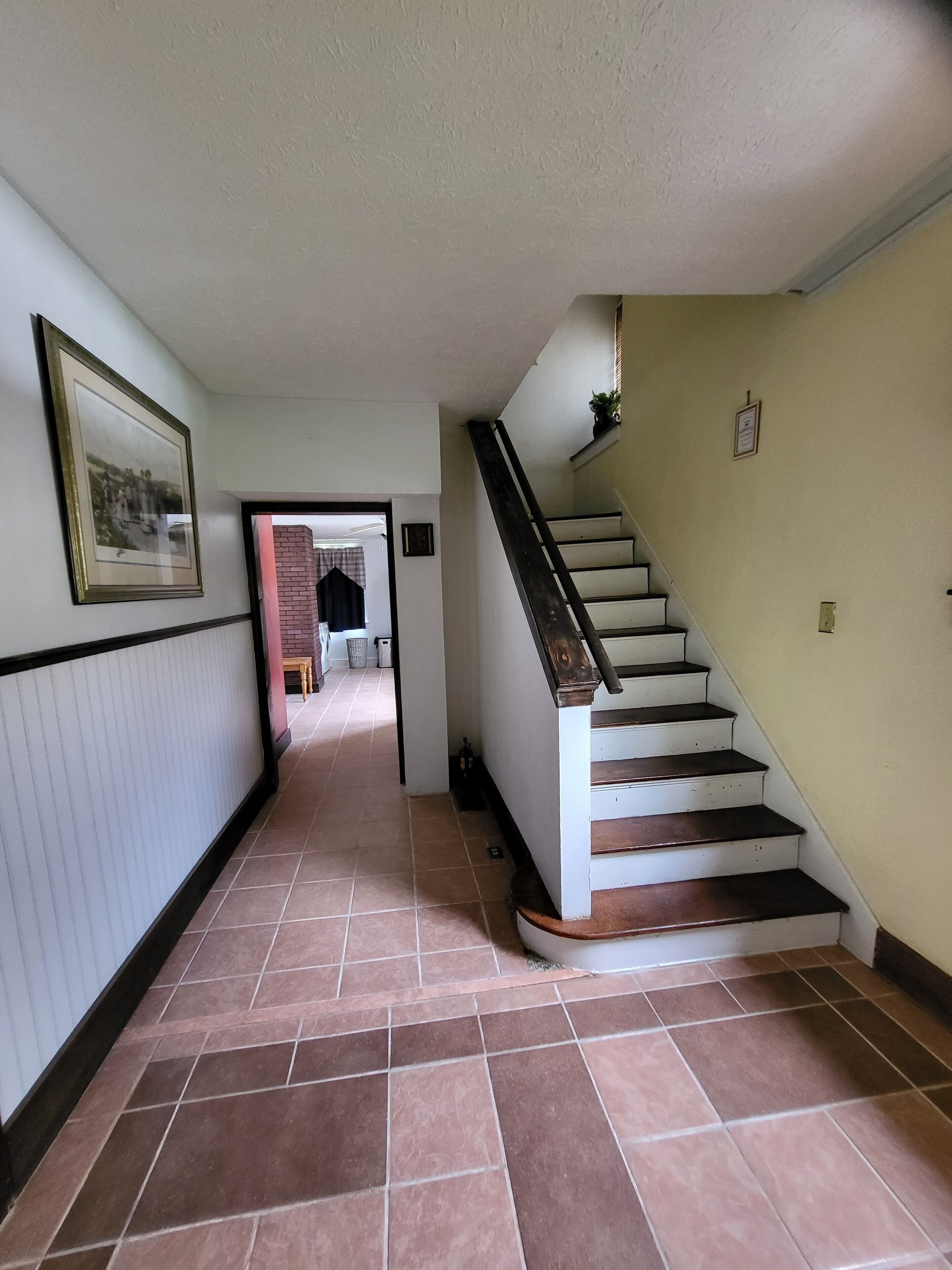 En hall med en trappa, ett klinkergolv och en inramad tavla på väggen.
