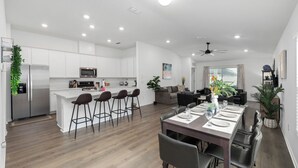 Una cocina moderna con armarios blancos, una isla central y taburetes de bar. Hay una zona de comedor con una mesa preparada para cuatro personas, y una sala de estar con un sofá y una mesa de centro.