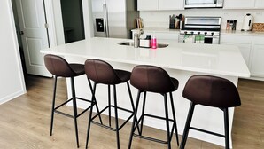 Una cocina moderna con armarios blancos, un refrigerador de acero inoxidable, horno empotrado y microondas. Hay cuatro taburetes de bar con asientos oscuros.