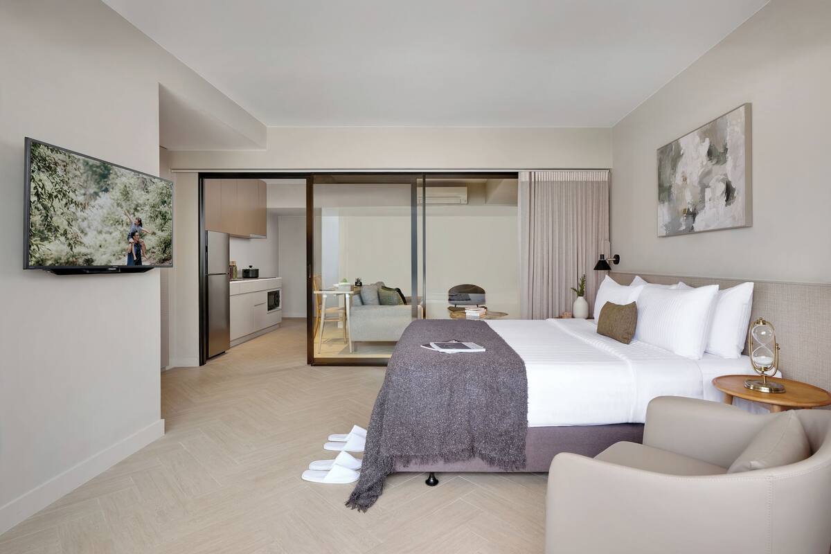 Una habitación de hotel moderna con una cama grande, un sofá, un televisor de pantalla plana, un área de comedor y un ventanal corredero que da a un balcón.