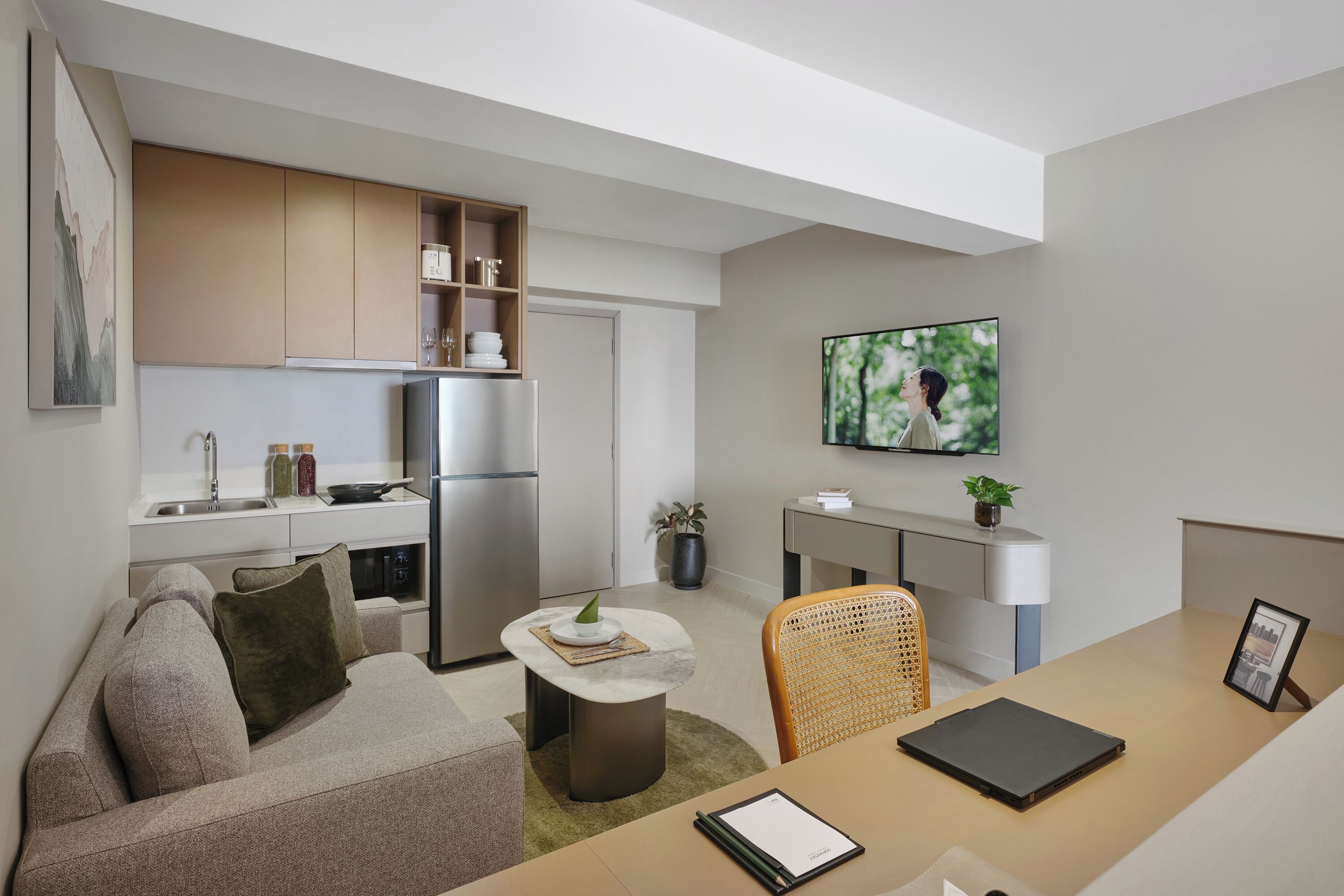 A modern living room with a sofa, a coffee table, and a TV. There is a kitchen area with a fridge, a sink, and cabinets.
