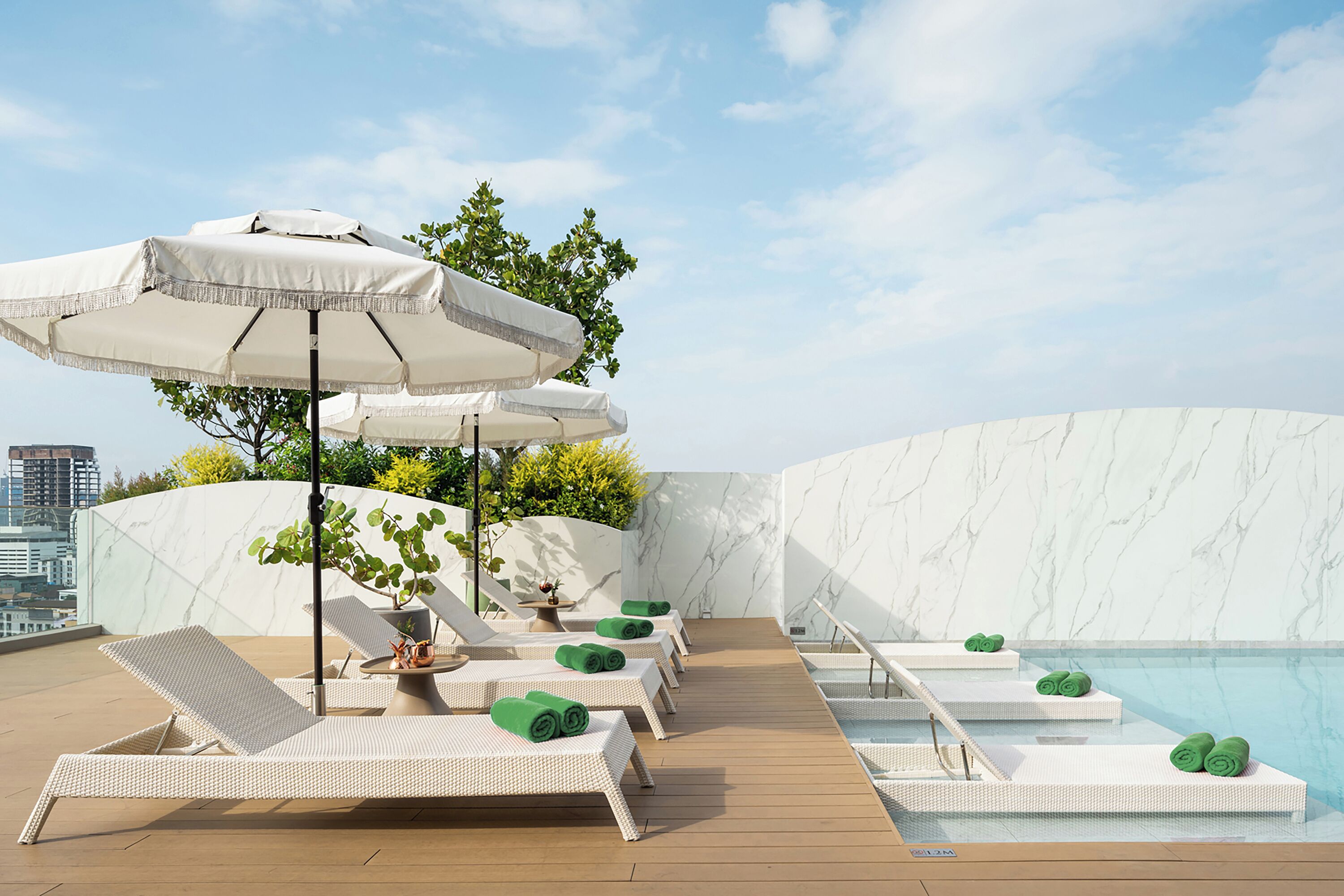 Un área de piscina en la azotea con sillones de descanso y sombrillas, una pared de mármol y vistas al horizonte de la ciudad.