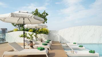 Un área de piscina en la azotea con sillones de descanso y sombrillas, una pared de mármol y vistas al horizonte de la ciudad.
