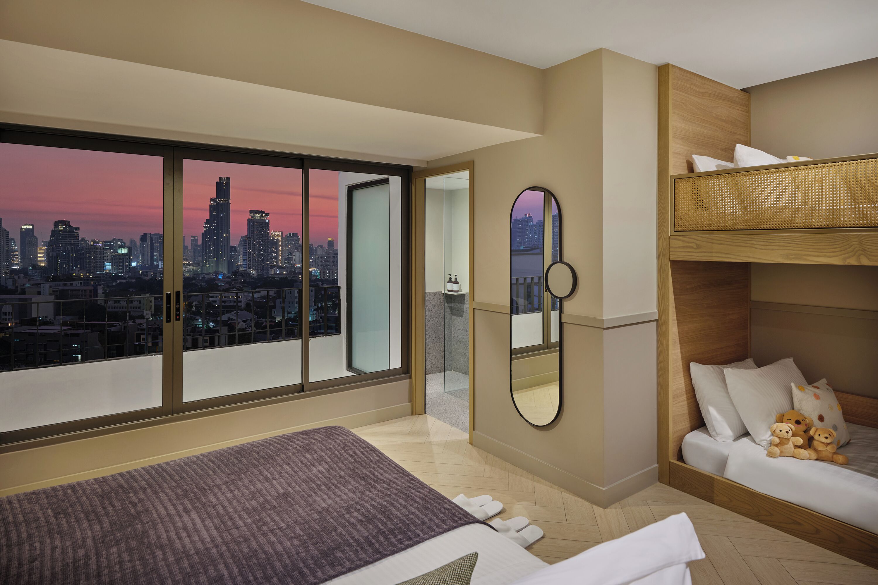 A modern hotel room with a city view, a large bed, a small sofa, and a wooden bunk bed.