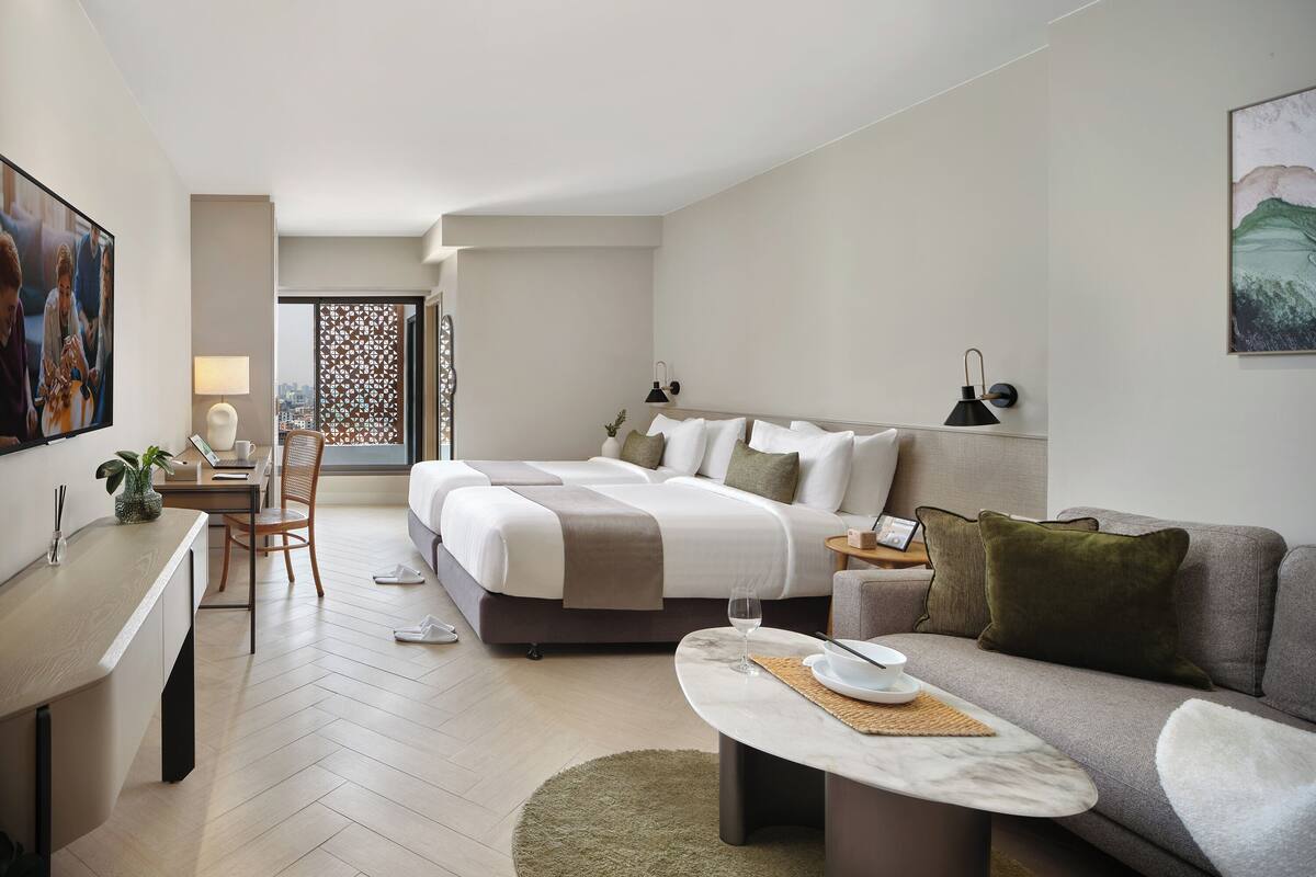 Una habitación de hotel moderna con una cama grande, un área de descanso con sofá y mesa de centro, un escritorio con silla y vistas a la ciudad desde la ventana.