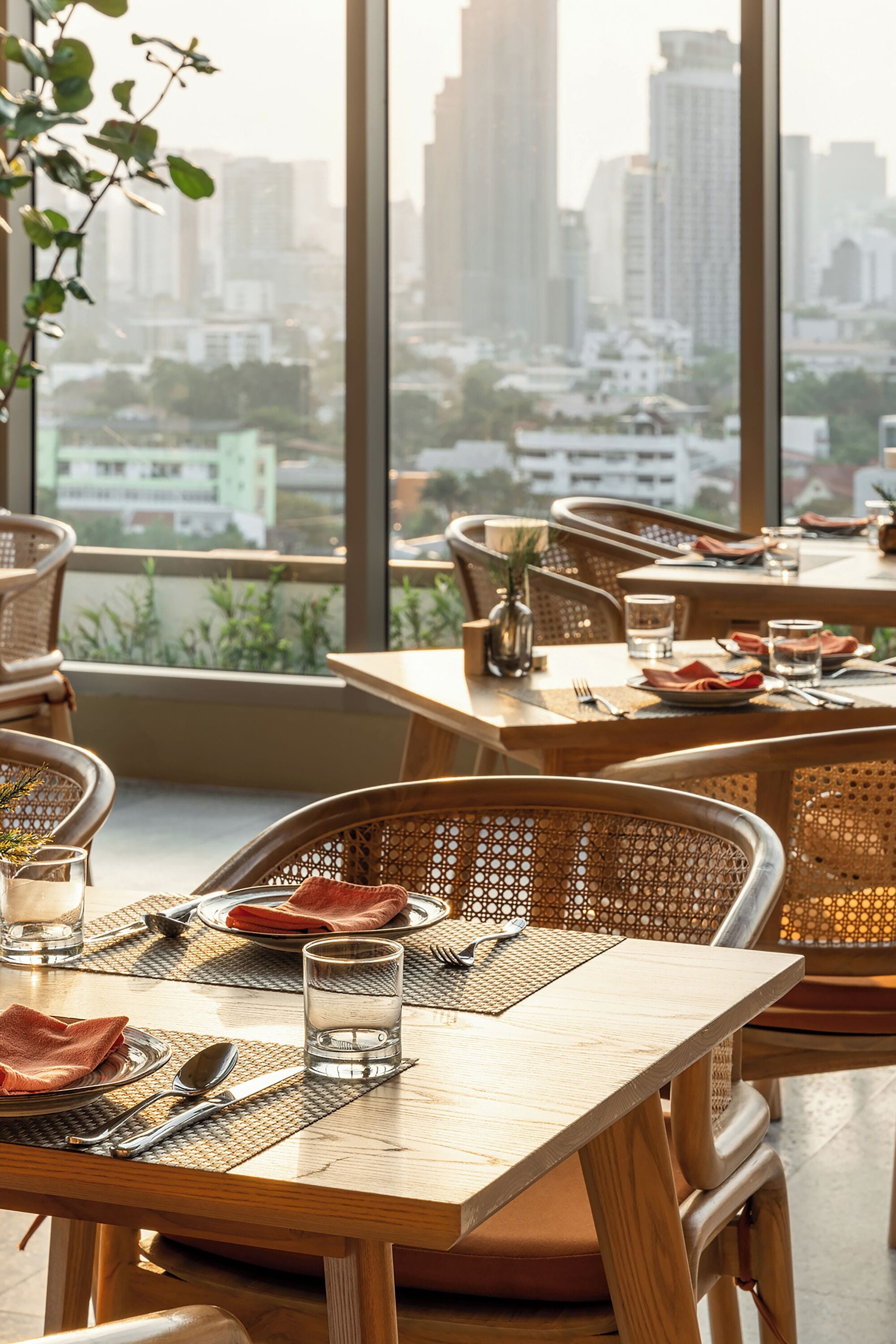 Comedor con mesas dispuestas para dos, con sillas de mimbre y vistas a un paisaje urbano a través de amplias ventanas.