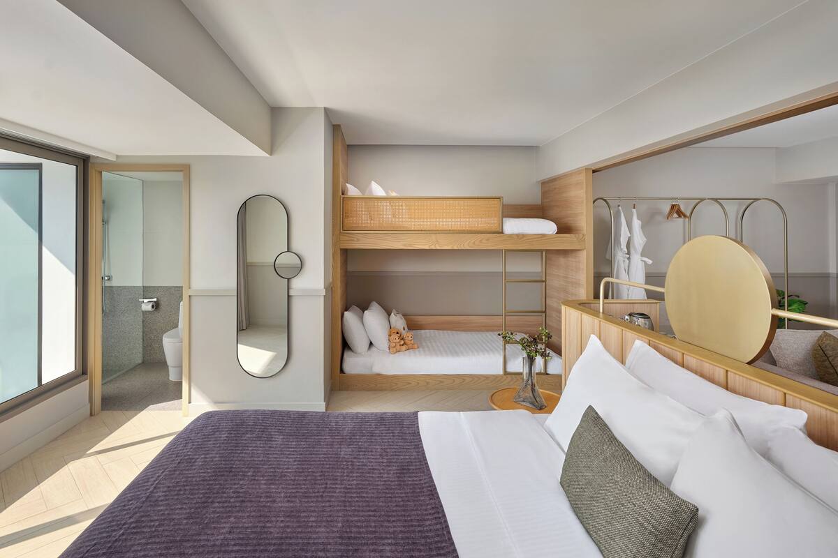 Una habitación de hotel moderna con una cama grande, una cama litera, un baño con puerta de cristal y un espejo grande.