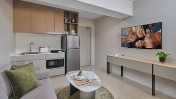 A modern kitchen with a built-in oven, microwave, and stainless steel refrigerator. There is a dining area with a marble table and a sofa with a green pillow. A flat-screen TV is mounted on the wall above the dining area.
