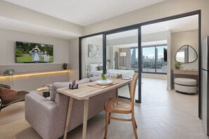 A modern living room with a sofa, dining table, and chairs. There is a television mounted on the wall, a plant, and a city view through large windows.