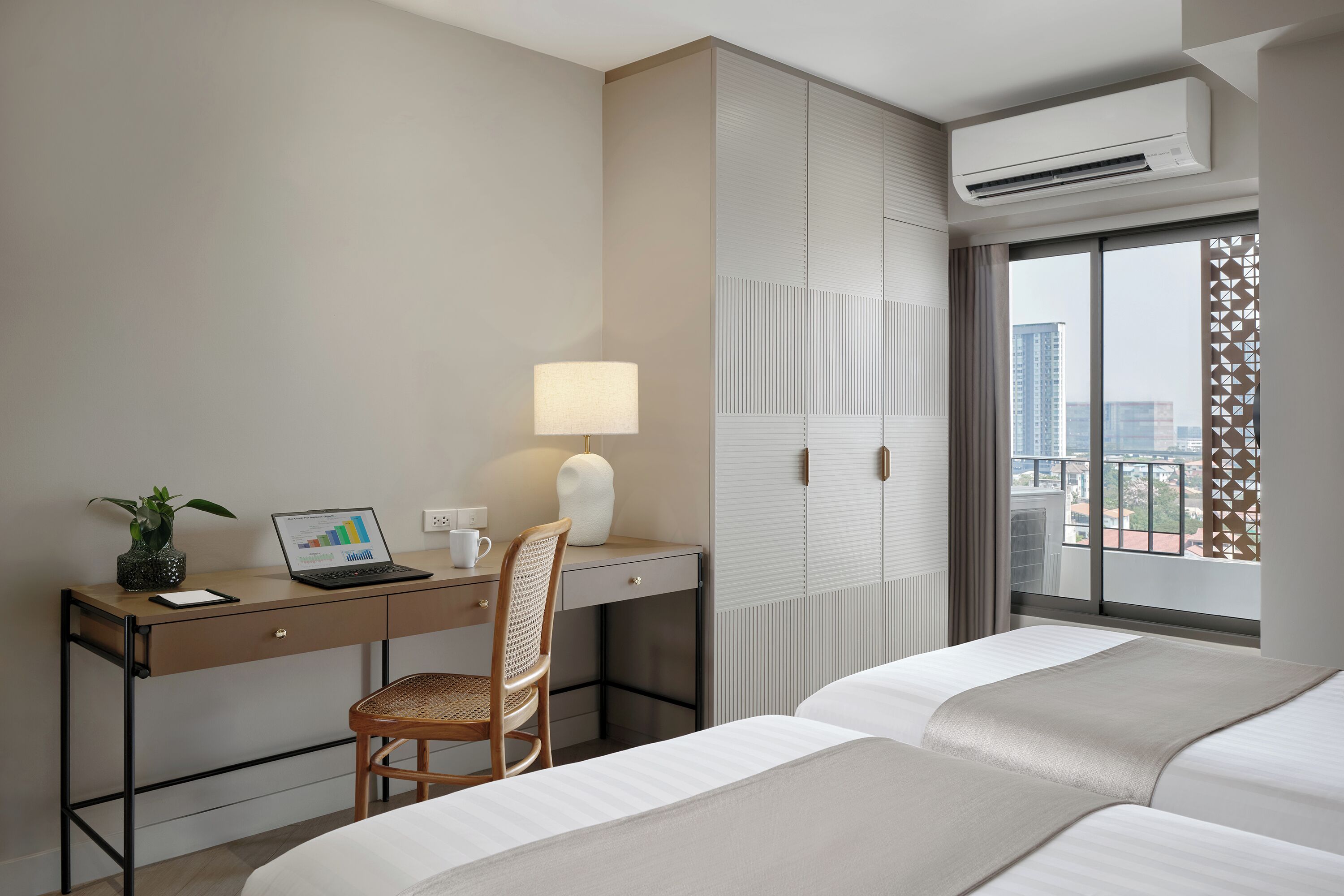 Ein Hotelzimmer mit einem Bett, einem Schreibtisch mit einem Laptop, einem Sessel und einer Lampe, sowie Blick auf die Stadt durch große Fenster.