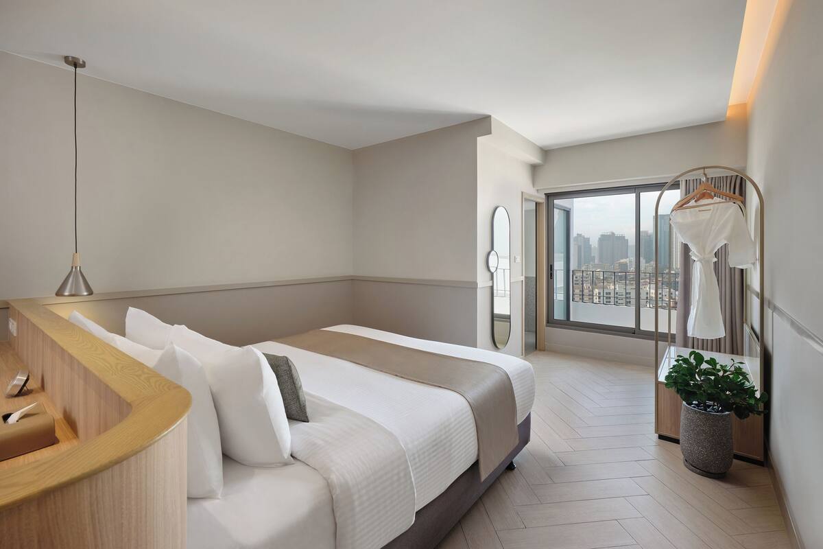 Una habitación de hotel moderna con una cama grande, una mesita de noche, un espejo, un perchero y una planta en maceta.