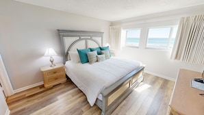 Un dormitorio con cama, mesita de noche, lámpara y vista al mar a través de las ventanas.