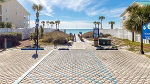 A beachside walkway with palm trees, a white fence, and a blue informational sign. - Grand Caribbean East 101 2 Bedroom Condo (Destin)