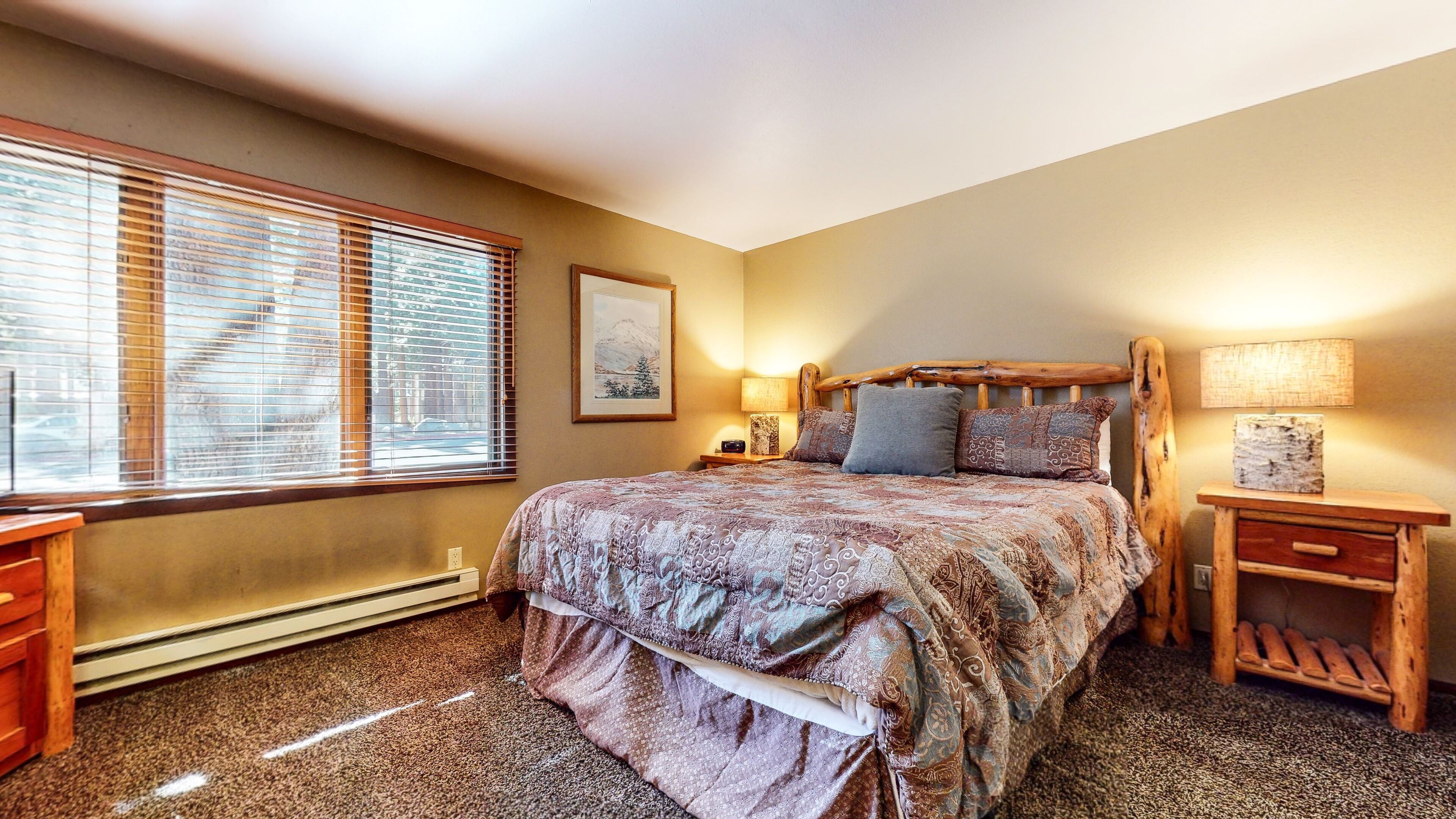 A bedroom with a large bed, two bedside tables, a heater, and a window with blinds.