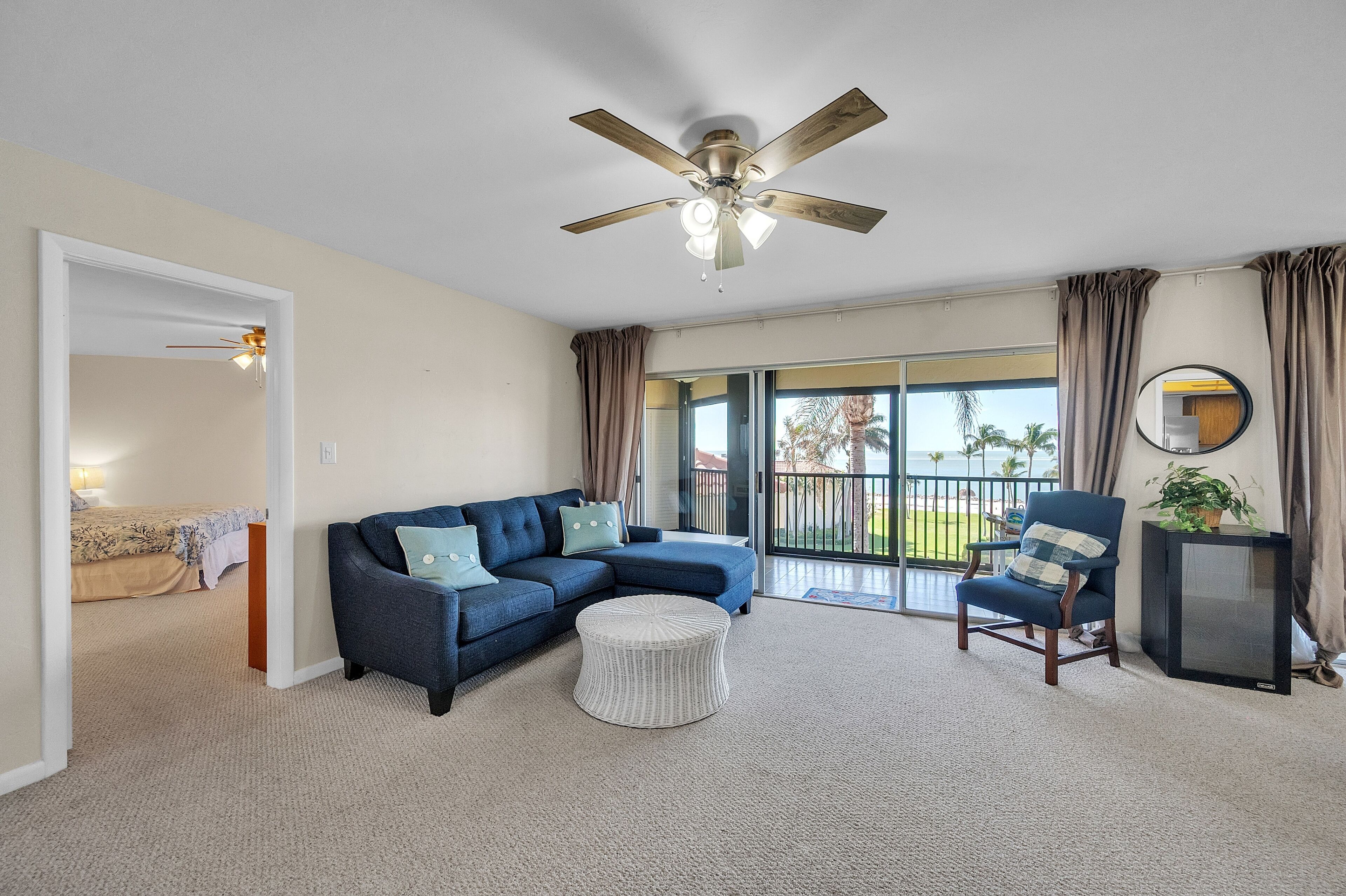A living room with a sectional sofa, a round ottoman, a ceiling fan, a television, and a sliding glass door leading to a balcony.