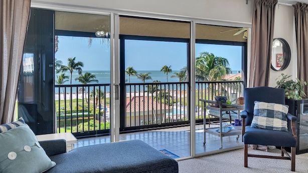 A balcony with a view of the ocean and palm trees, a chair with a checkered pillow, and a table with a plant. - Gulf-view Condo With Resort-style Amenities 2 Bedroom Condo (Marco Island)
