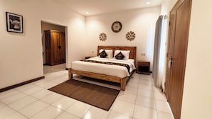 A bedroom with a wooden bed, white bedding, a small bedside table, a nightstand, a door, and a wall-mounted flower decoration. - Lubdhaka Canggu (Canggu)