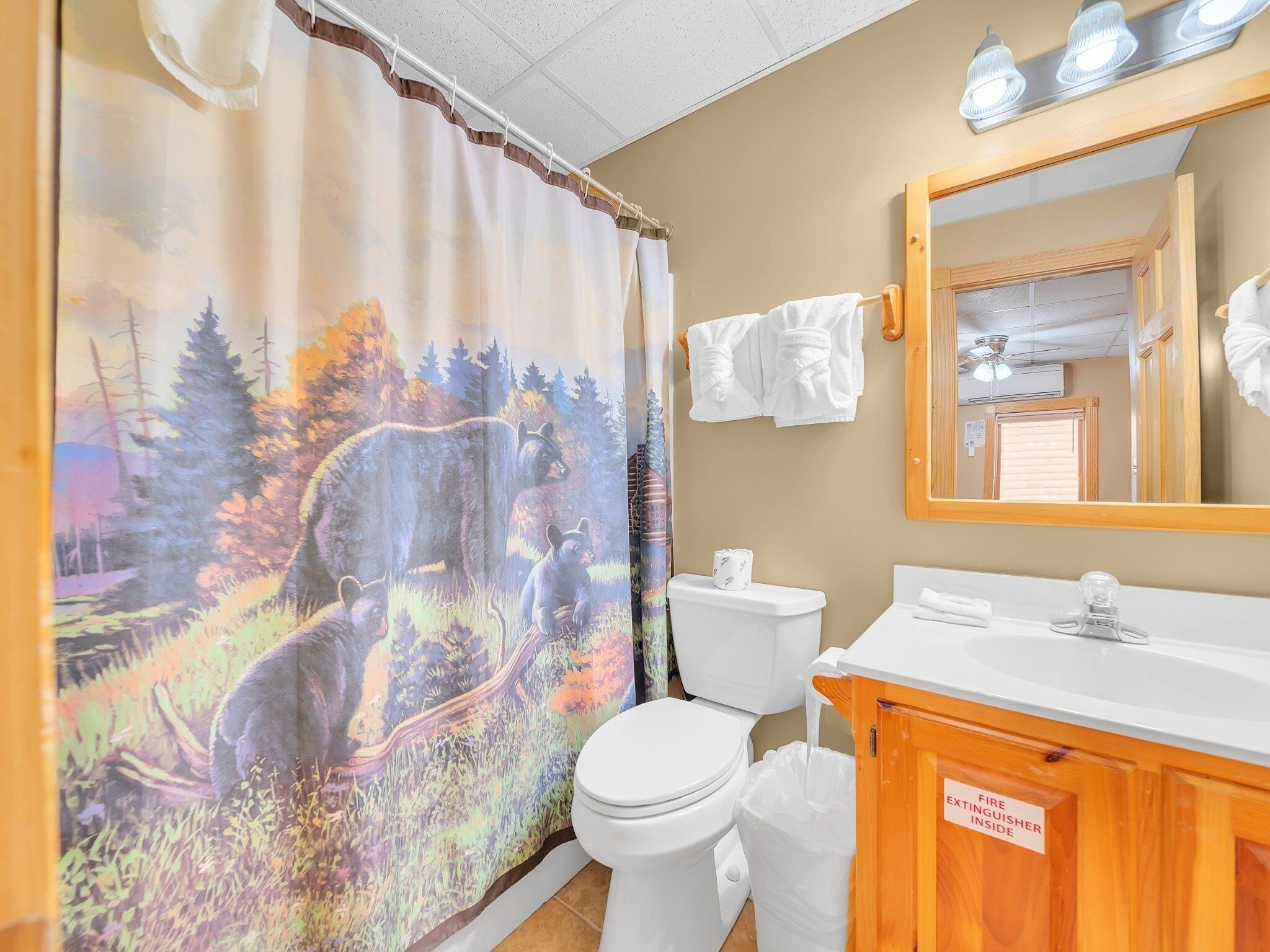 A bathroom with a forest-themed shower curtain, a white toilet, a sink with a mirror, and a fire extinguisher sign.