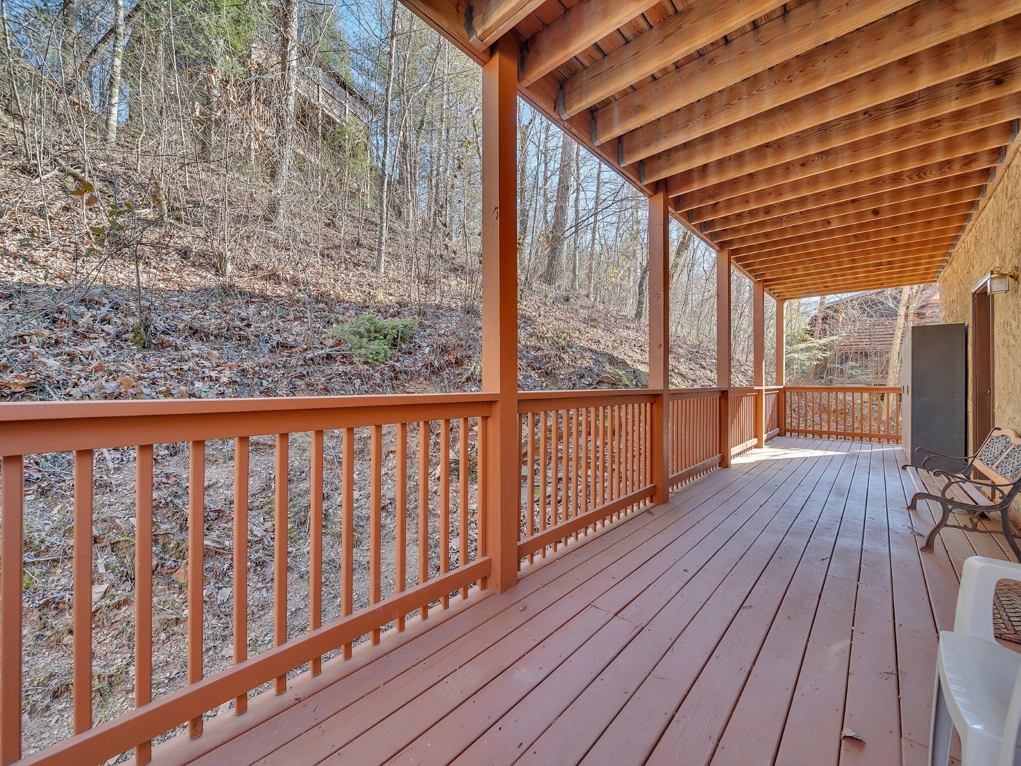A covered wooden deck with a railing, benches, and a view of a forested area.