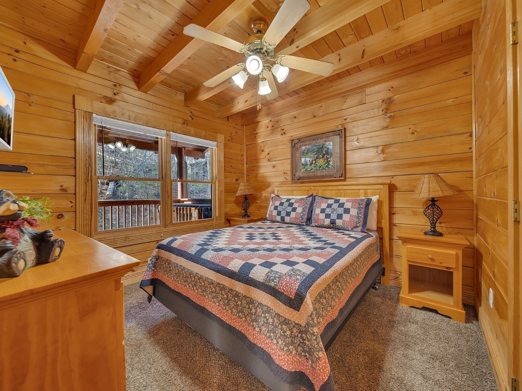 A cozy cabin bedroom with a large bed, wooden walls, a ceiling fan, and a window overlooking a deck.