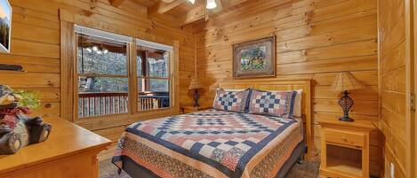 A cozy cabin bedroom with a large bed, wooden walls, a ceiling fan, and a window overlooking a deck.