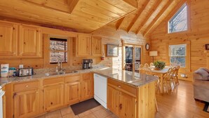 Una cabaña de madera con cocina que incluye armarios de madera, encimera de granito y una isla grande. Hay una zona de comedor con mesa y sillas, y vistas al exterior a través de las ventanas.