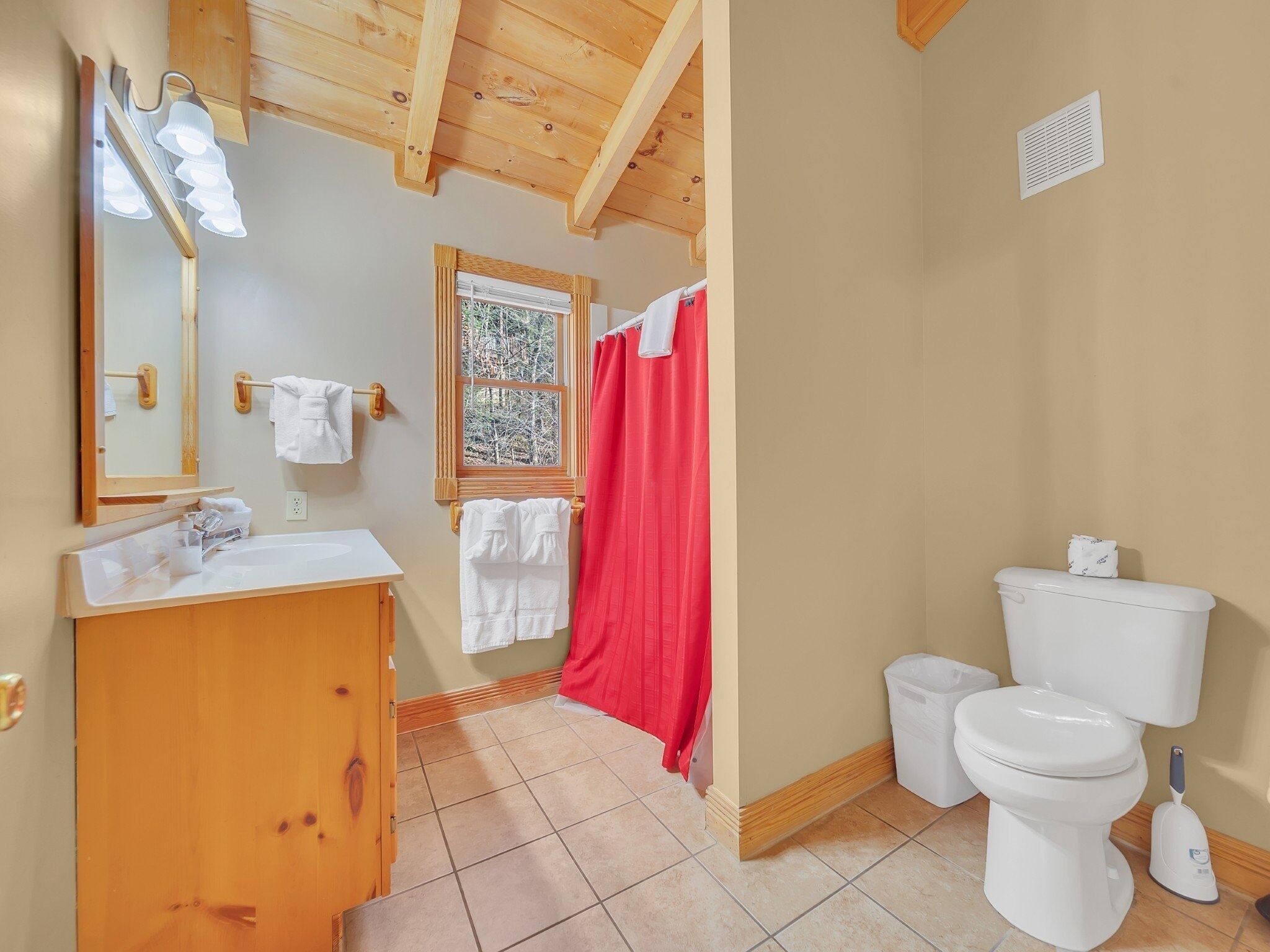 A bathroom with a wooden vanity, a toilet, a shower curtain, and a window with a view of trees.