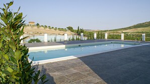 A rectangular swimming pool with a tiled deck, surrounded by a metal fence, set against a hilly landscape.