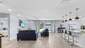 A modern kitchen with a bar area, a dining space, and a living area with sofas and a TV. - Soak Up The Sun - Saltwater Pool, Spacious Fenced-in Backyard, 3 Bedroom Home (Cape Coral)