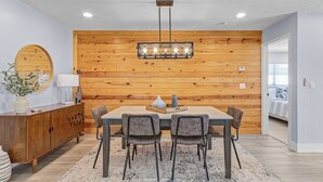 A dining area with a wooden wall, a hanging light fixture, a wooden cabinet, and a table with chairs. - Soak Up The Sun - Saltwater Pool, Spacious Fenced-in Backyard, 3 Bedroom Home (Cape Coral)