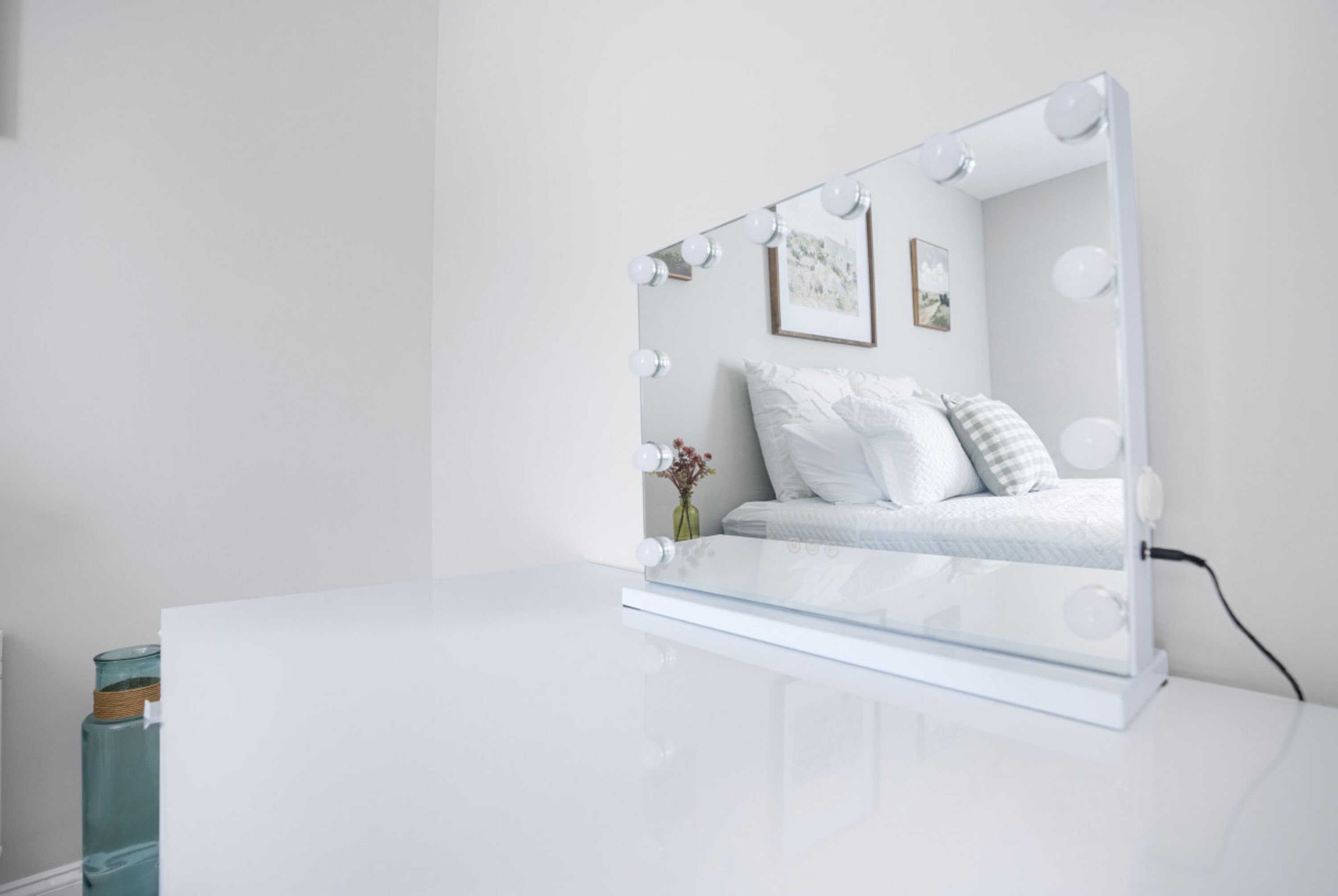A bedroom with a vanity mirror, bed, pillows, and a vase with flowers.